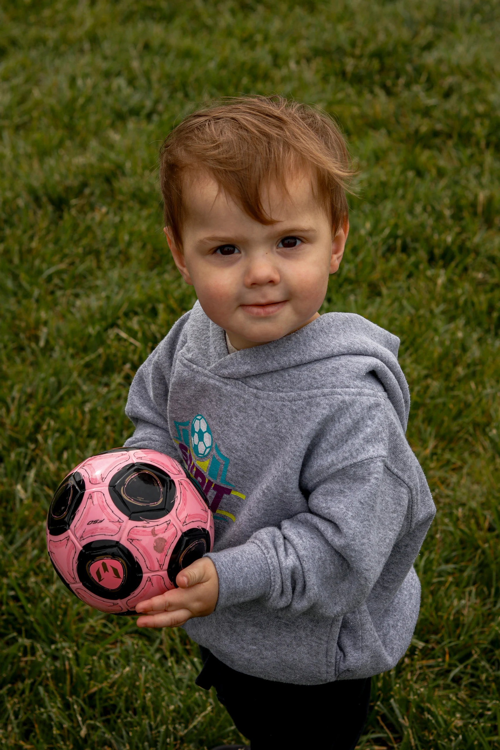 A young child looks toward the camera while holding a pink soccer ball.