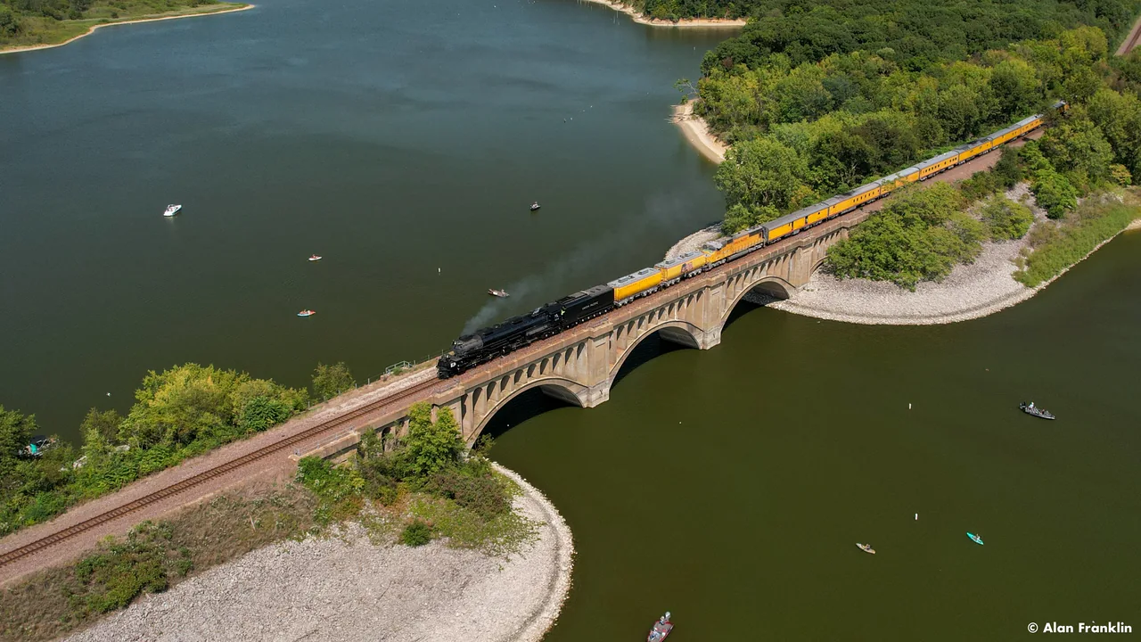 Big Boy and its passenger cars cross an arched bridge over a lake.