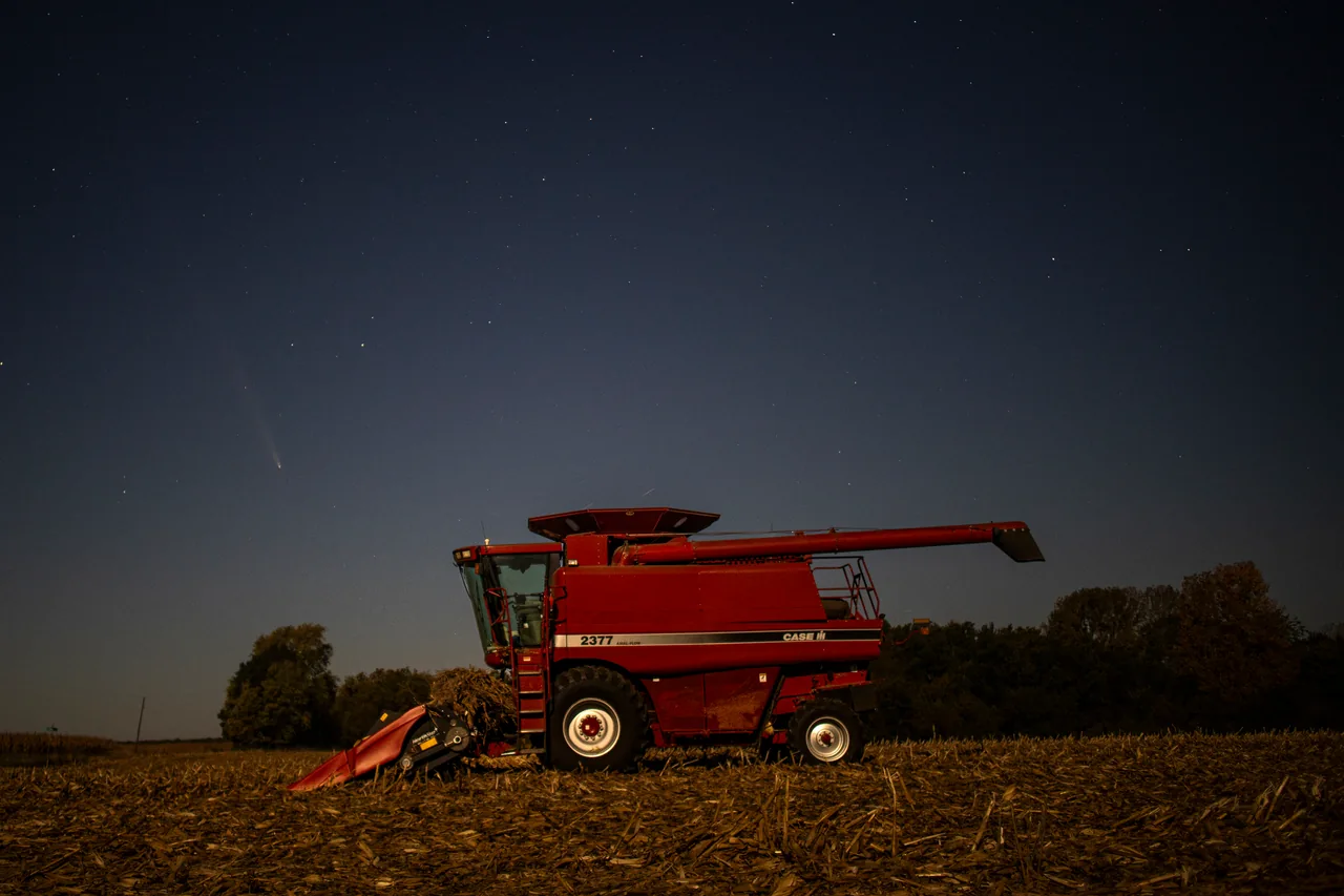 A comet above a red combine parked in a harvested field at night.