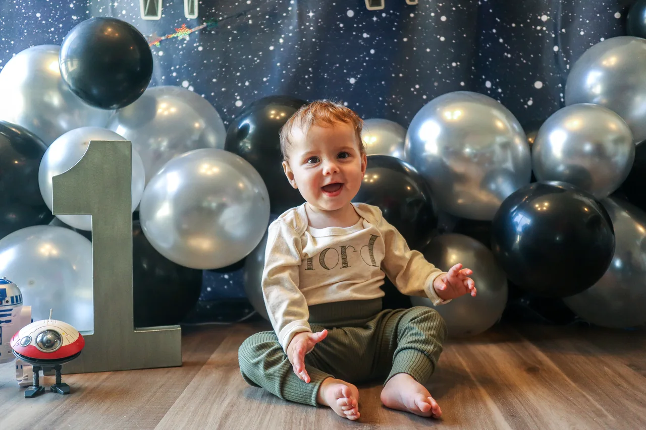 A smiling child sits among silver and black balloons at a first birthday celebration.